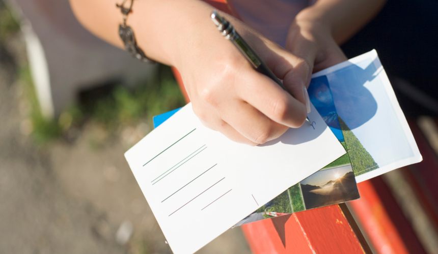 Close-up of a hand writing a message on the back of a postcard with a pen.