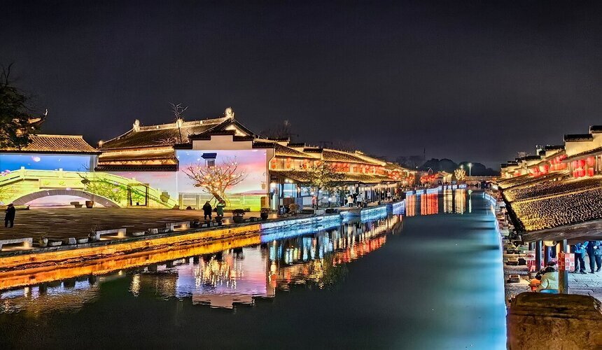 People walking along a riverside promenade in China at night with city lights reflected on the water.